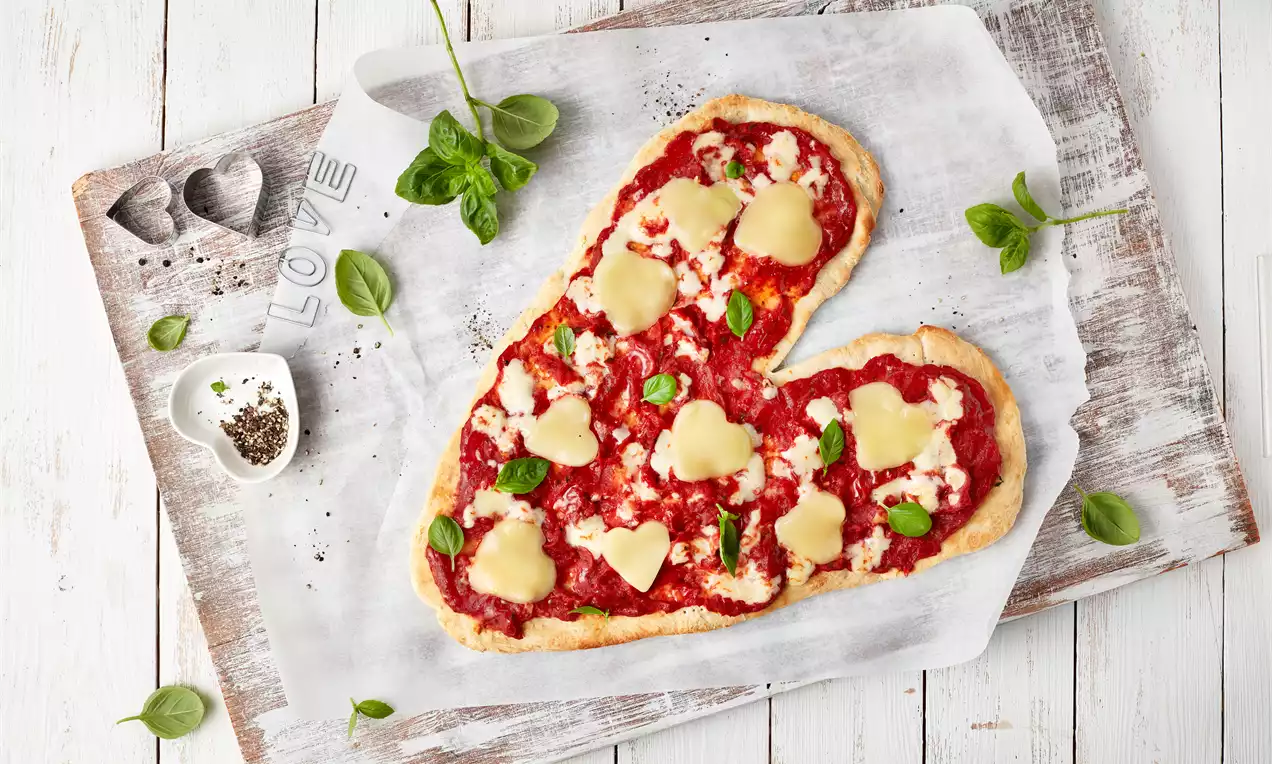 Pizza u obliku srca na papiru za pečenje, prekrivena crvenim sosom, zelenim listićima i trakama sira te dekorirana srcima od sira