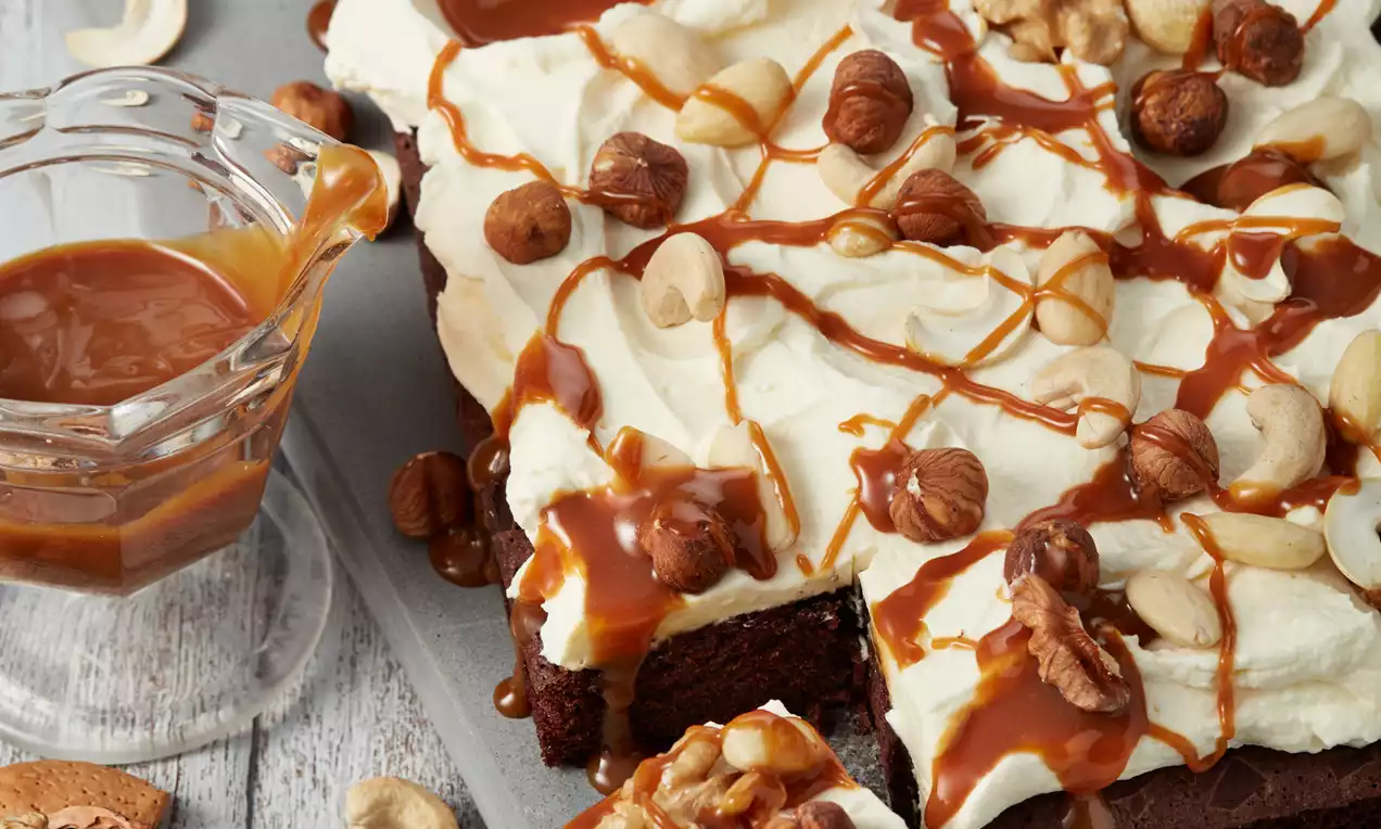 Brownies tamnosmeđe boje prekriven kremom od sira, ukrašeni karamel preljevom i orašastim plodovima na sivom pladnju