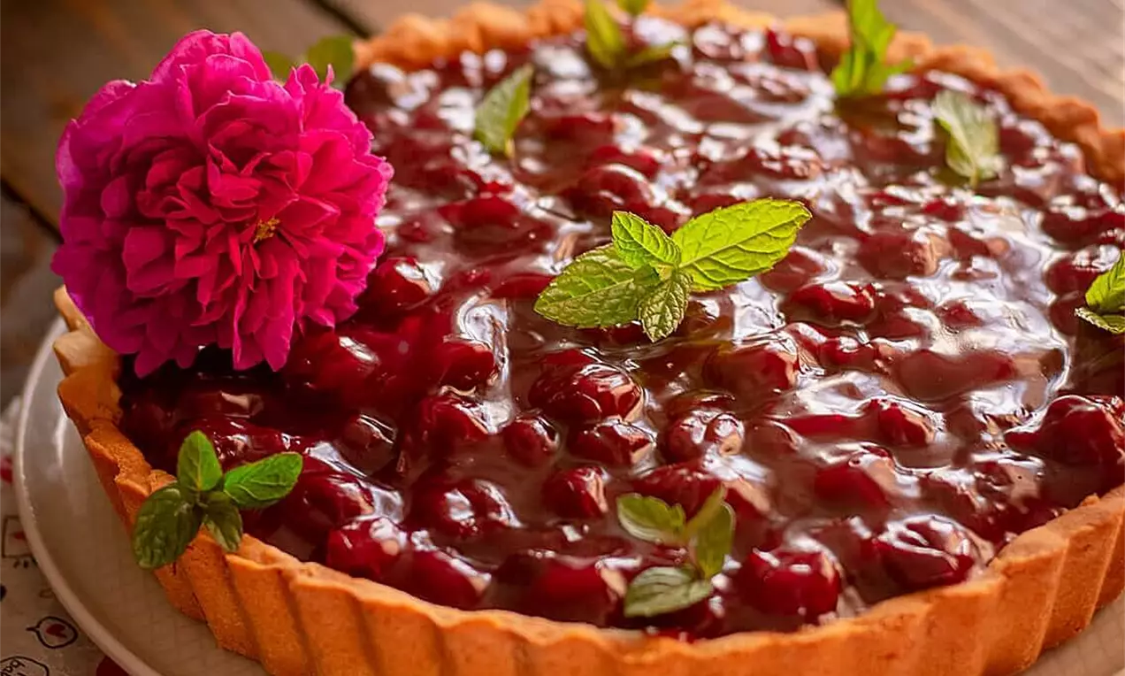 Tart s hrskavom, rebrastom podlogom zlatno smeđe boje, sjajnim preljevom od crvenih trešanja, ukrašen listićima mente i ružičastim cvijetom na rustičnom drvenom stolu