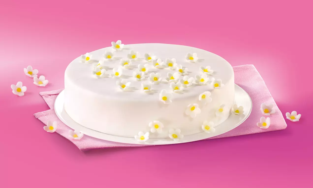 Okrugla torta s glatkom bijelom glazurom posuta malim bijelim cvjetovima sa žutim središtem, na bijelom tanjiru i ružičastoj podlozi