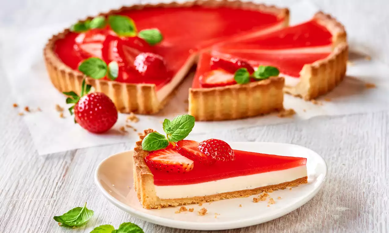 Tart s hrskavom zlatnom koricom, glatkim slojem bijele panna cotte i svijetlocrvenim preljevom od jagoda, ukrašen svježim listićima mente