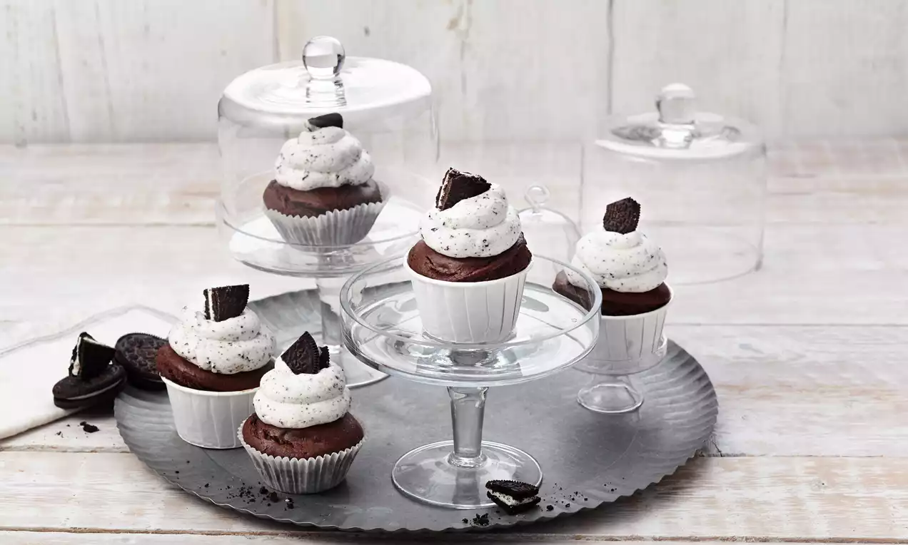 Čokoladni cupcakes u bijelim košaricama s bijelom kremom prošaranom crnim mrvicama, ukrašeni komadićima Oreo keksa na metalnom poslužavniku