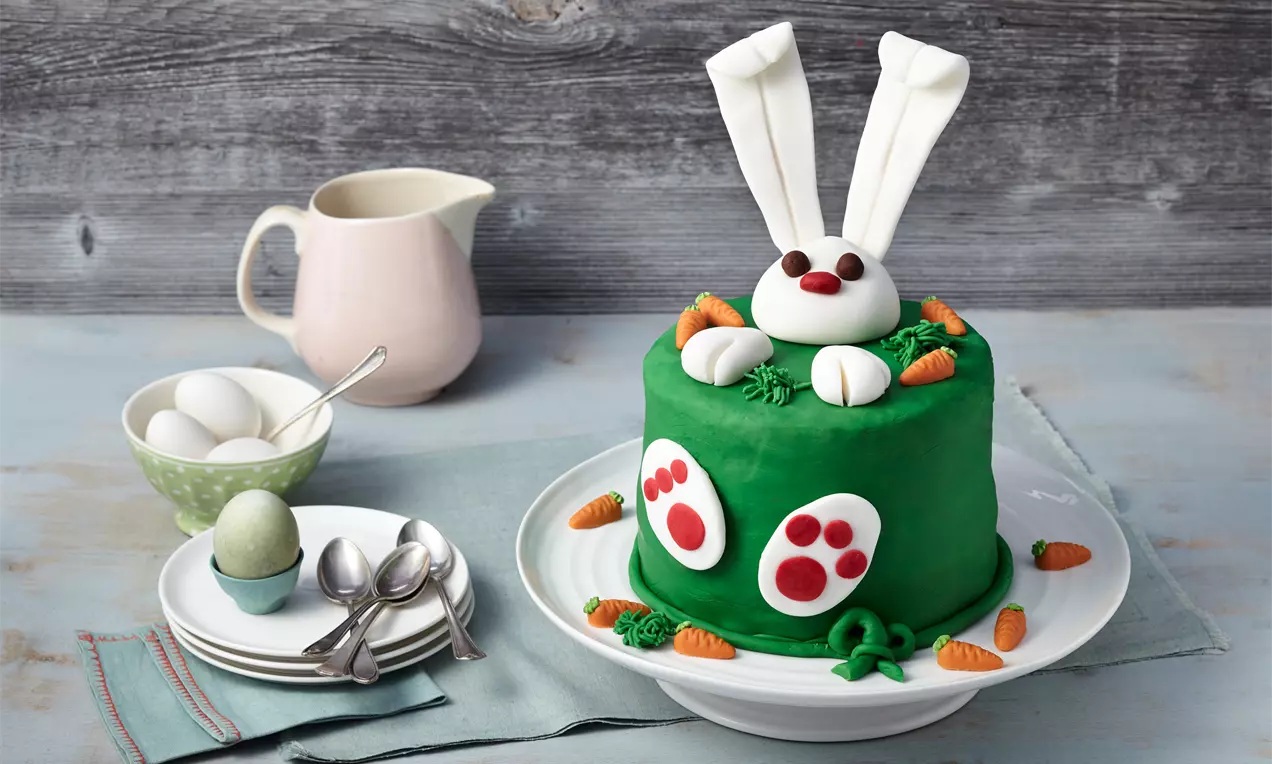 Okrugla torta presvučena zelenom glazurom ukrašena fondant figurama zeca s bijelim ušima, šapama i narančastim mrkvama na bijelom tanjiru