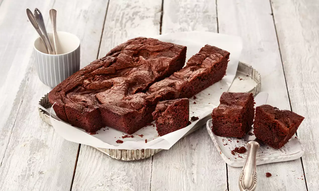 Sočni tamnosmeđi brownie s lagano ispucalom koricom, izrezan na kocke, poslužen na bijelom papiru u rebrastoj metalnoj posudi.