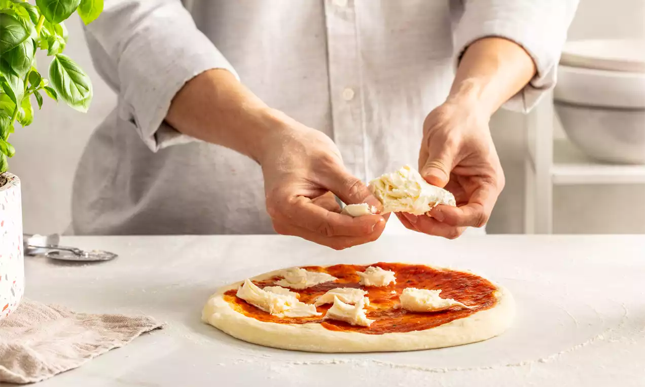 Pizza od kiselog tijesta svijetložute boje, s crvenim slojem umaka, nepravilno raspoređenim komadićima sira na bijelom radnom stolu