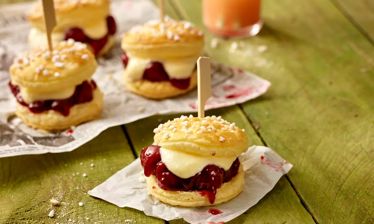 Mini deserti od zlatnopečenog lisnatog tijesta punjeni tamnocrvenim voćnim nadevom i svijetlom kremom, posuti krupnim šećerom na papiru