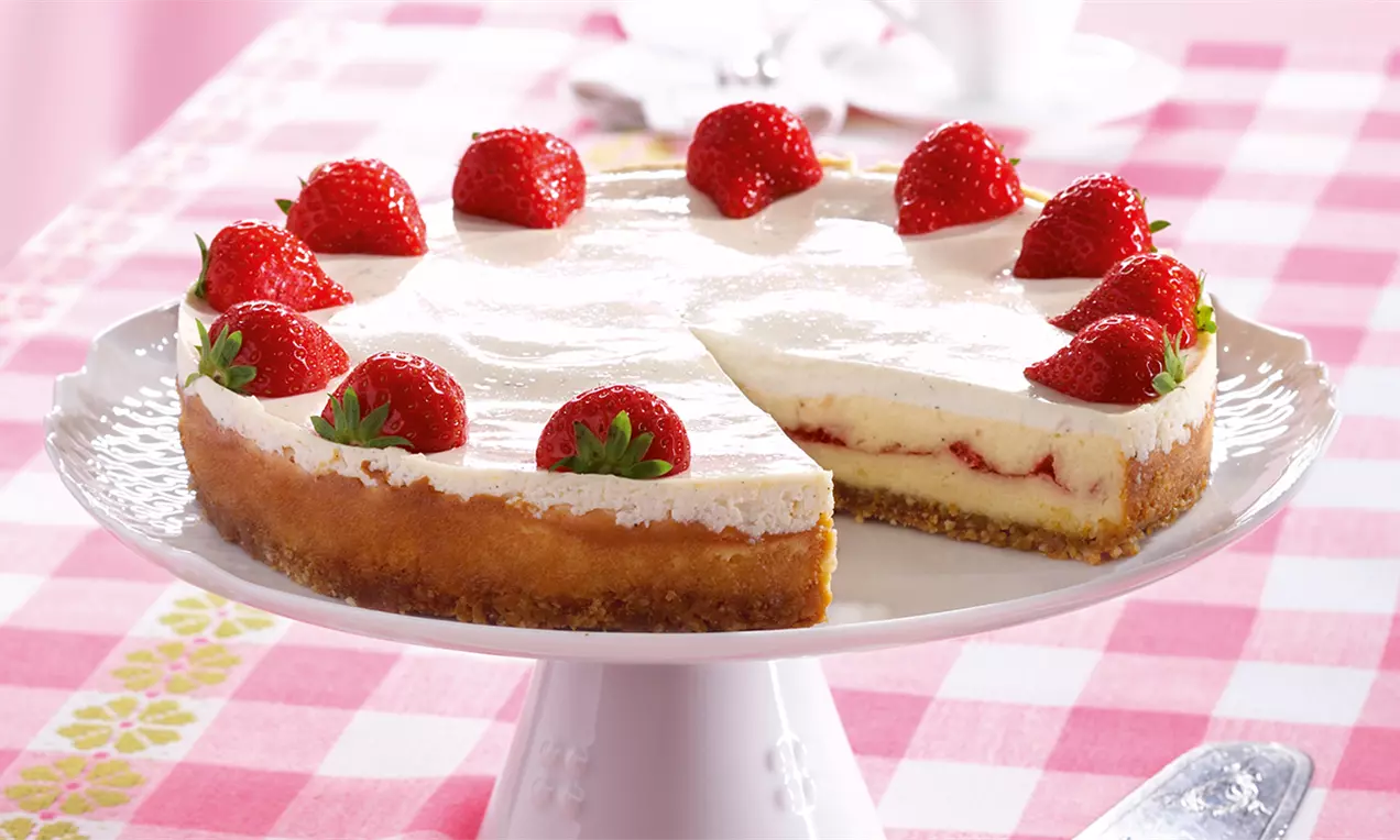 Okrugla torta s hrskavom smeđom podlogom, bijelim krem slojem i cijelim crvenim jagodama na vrhu, na bijelom stalku za torte