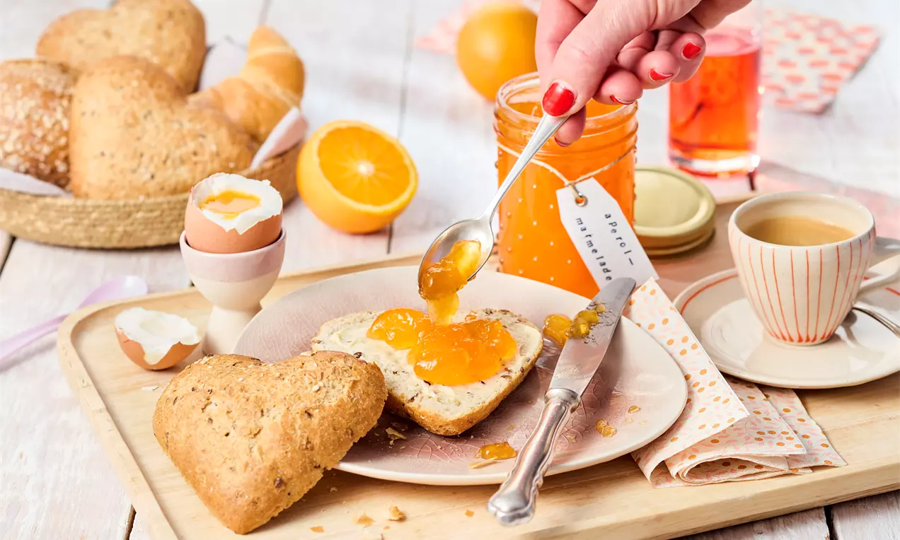 Svijetlonarančasta marmelada razmazuje se na svijetlo smeđoj kruškoj polovici, okružena jajima, pecivima, kriškama naranče i šoljicom kafe