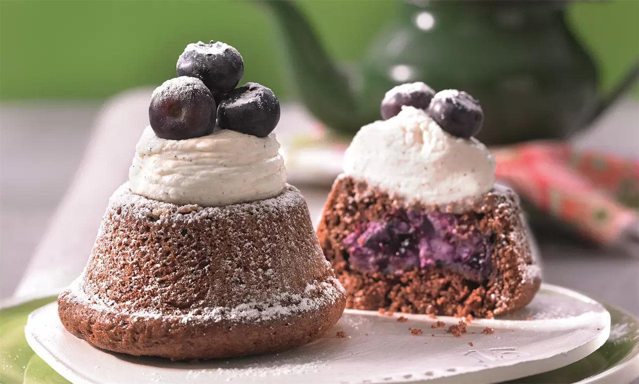 Mini tortice od tamnog biskvita s vrhom od bijele kreme, posute šećerom u prahu, ukrašene borovnicama, jedna prerezana otkriva ljubičasti nadjev