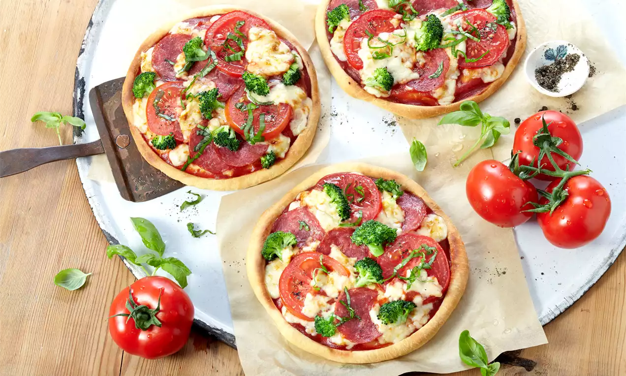 Mini pizze s tankom, zlatnosmeđom korom, slojevima rastopljenog sira, kriškama crvenog paradajza, kobasice i svježe brokule na papiru za pečenje