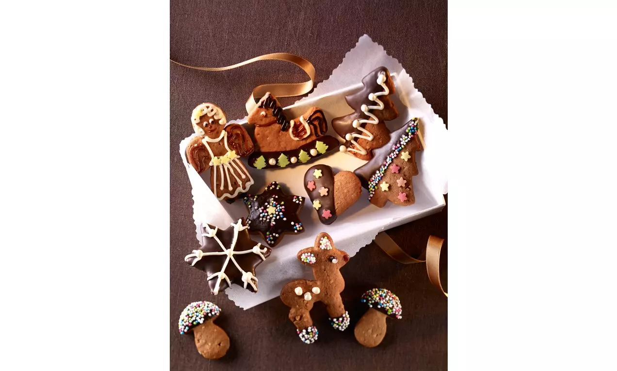 Raznovrsni lebkuchen kolačići tamnosmeđe boje, ukrašeni šarenim glazurama i mrvicama, složeni na bijelom papiru sa zlatnom vrpcom